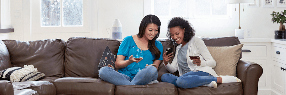 Two females on couch holding wine glass & looking at cell phone