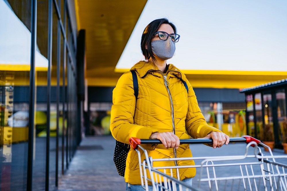 Short-haired woman in a yellow jacket wearing a mask pushes a shopping cart out of a grocery store.
