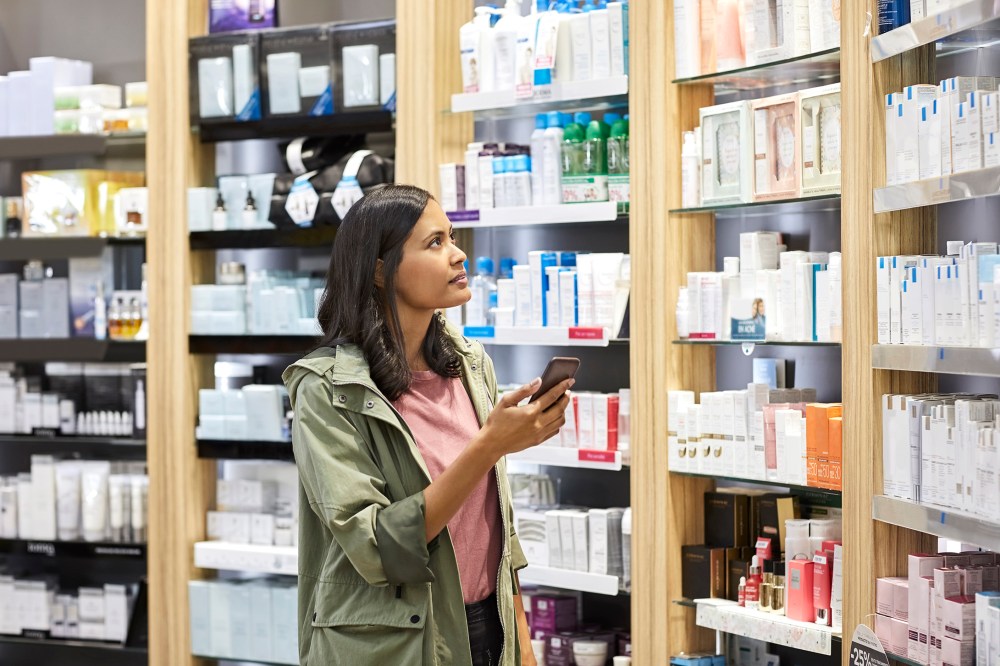 Female customer searching medicine on shelf from rack at pharmacy. Young woman is holding smart phone.