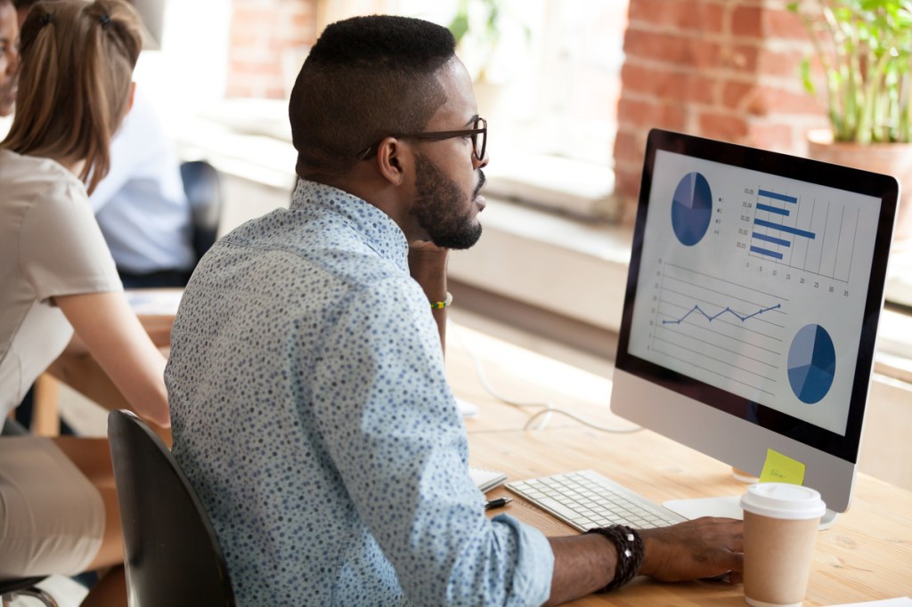 Serious African American man in glasses talking by phone about economic report with pie diagrams, marketing plan, business results, using computer, applications, looking at screen