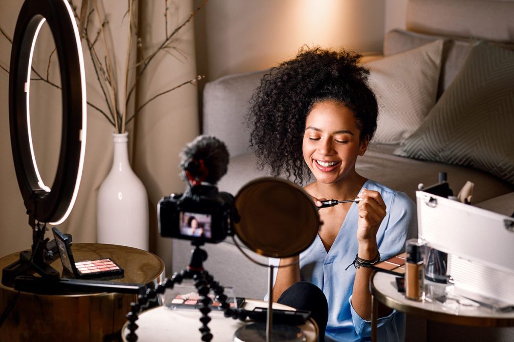 A black woman applies makeup in front of a camera, mirror and ring light.