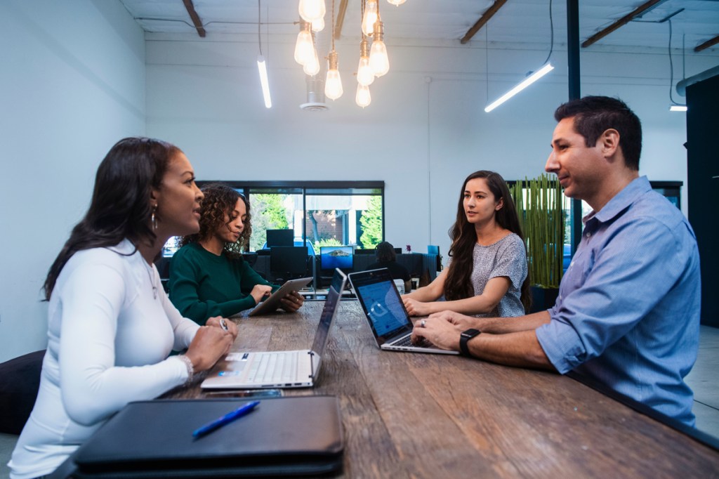 Bright conference room with 4 people conversing with laptops