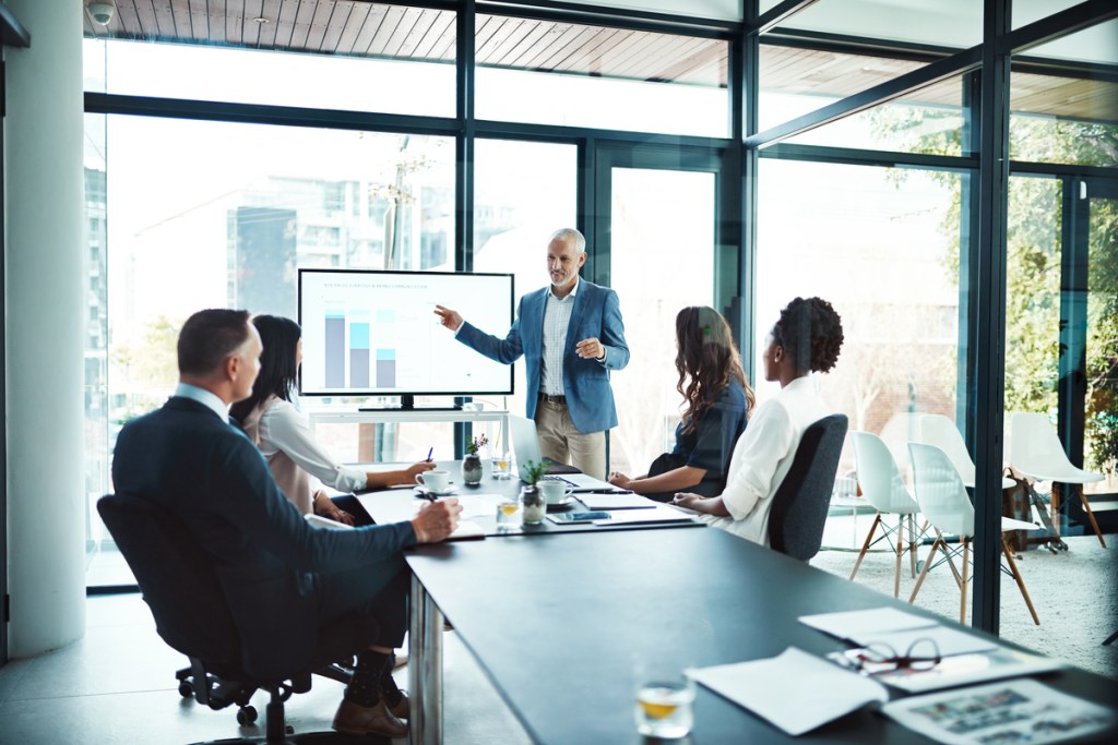 Large board room with man presenting in front of a screen to 4 team members