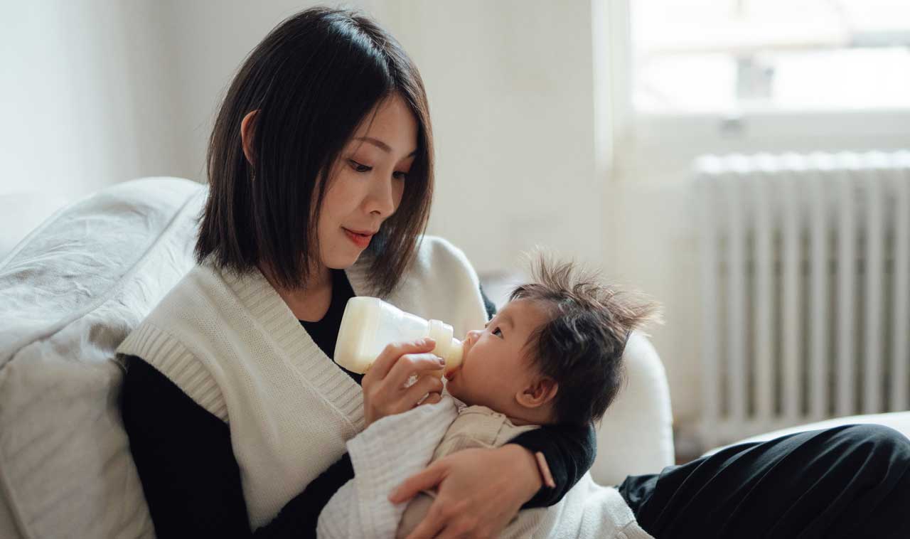 Asian woman with white sweater vest bottle feeds her child