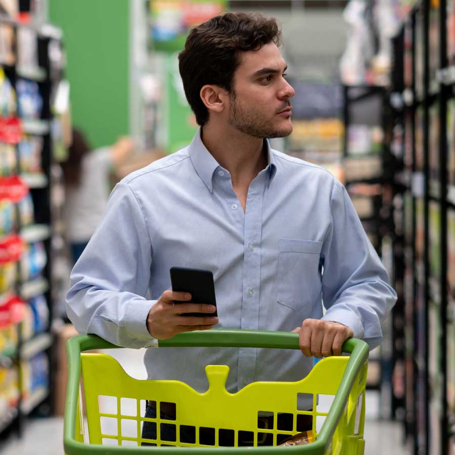 Man with white collared shirt looking at products in a store holding a mobile phone and pushing a green shopping cart