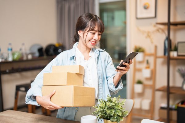 A young Asian American consumer is sitting at home, using her credit card and phone/laptop to make an online shopping transaction.