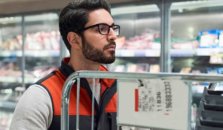 Man with dark hair and black framed glasses with an orange and black fleece in the dairy aisle of a grocery store