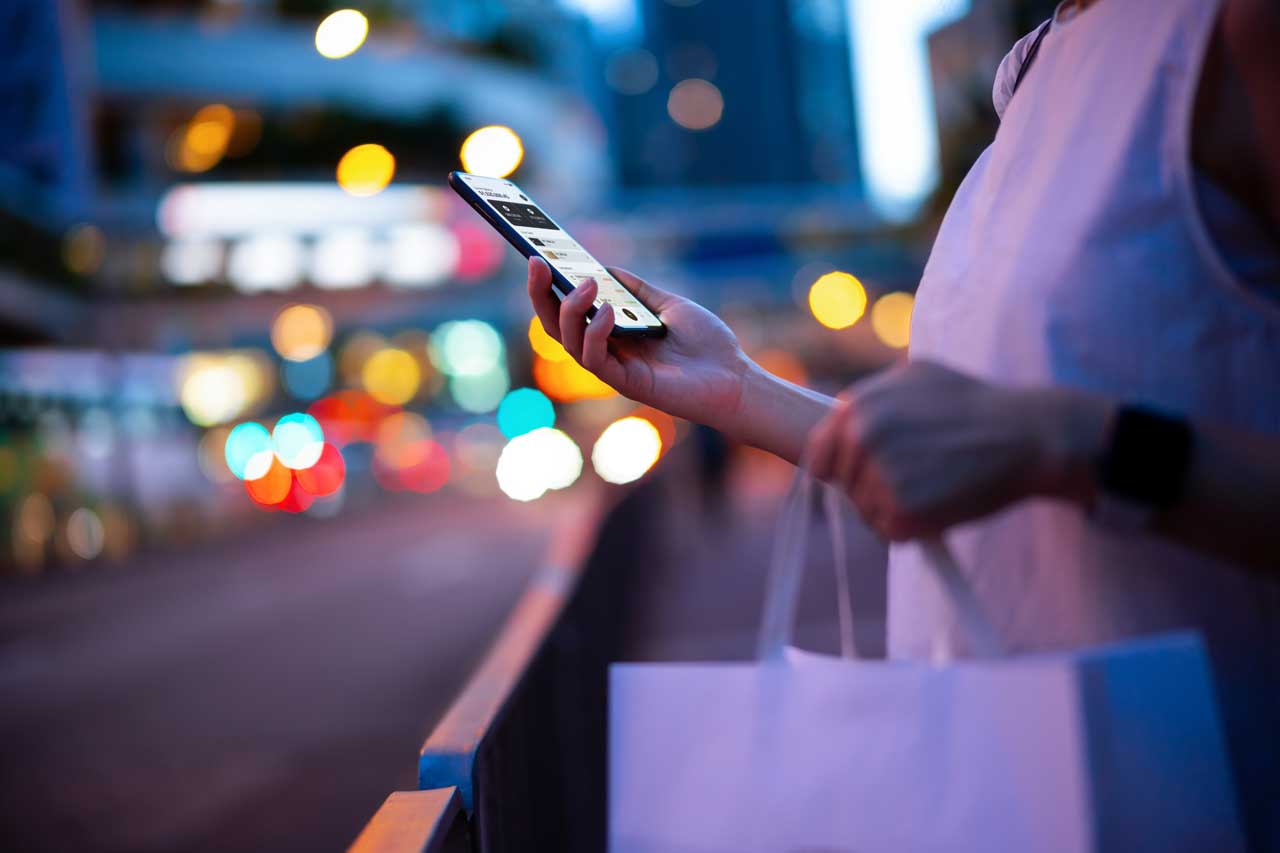 Closeup of woman holding a shopping bag and looking at her mobile phone on a busy street at night. 