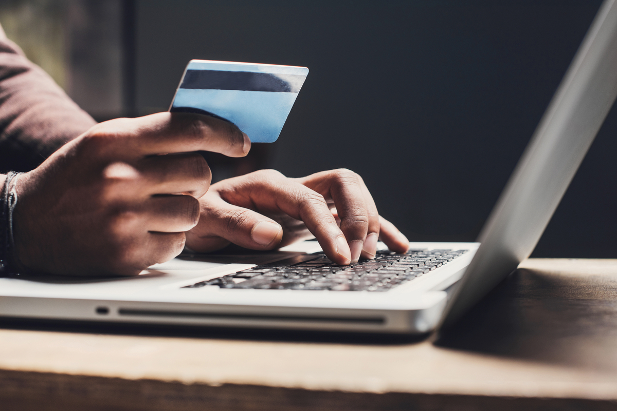 A man holds a credit card in one hand, while typing on a laptop, implying he's shopping online.
