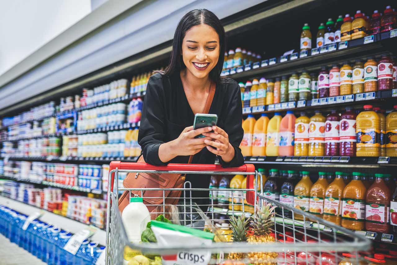 Woman with dark hair smiles as she looks at her phone while pushing a shopping cart in a store