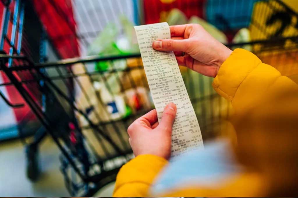 Woman in yellow coat holding a long receipt in front of a shopping cart