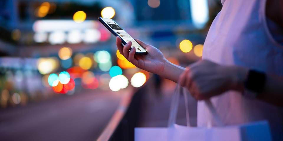Woman holding white shopping bag in a busy night setting looking at her phone
