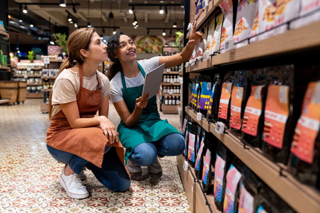 2 female store associates crouch at a bottom shelf. One points to the price and the other looks pensively.