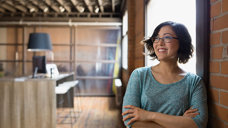 Asian woman with glasses standing near wall with arms crossed in open room
