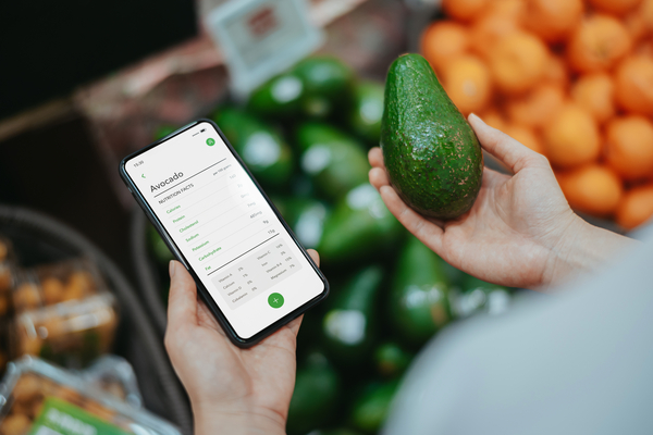 Asian woman shopping for fresh organic fruits and vegetables in supermarket. She is holding an avocado, using health and fitness tracker app on smartphone to check the nutrition facts and calories intake. Healthy eating lifestyle. Lifestyle and technology