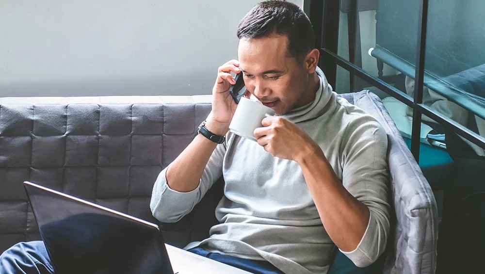 Asian man in grey shirt on phone drinking cofee out of a white mug while looking at a laptop