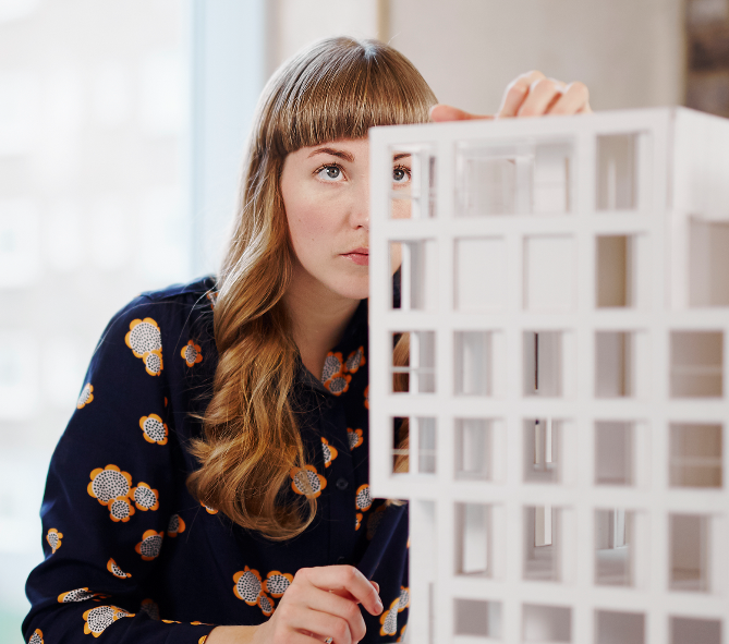 Woman with crew cut blond hair wearing a flower pattern shirt looking at a model of a white building