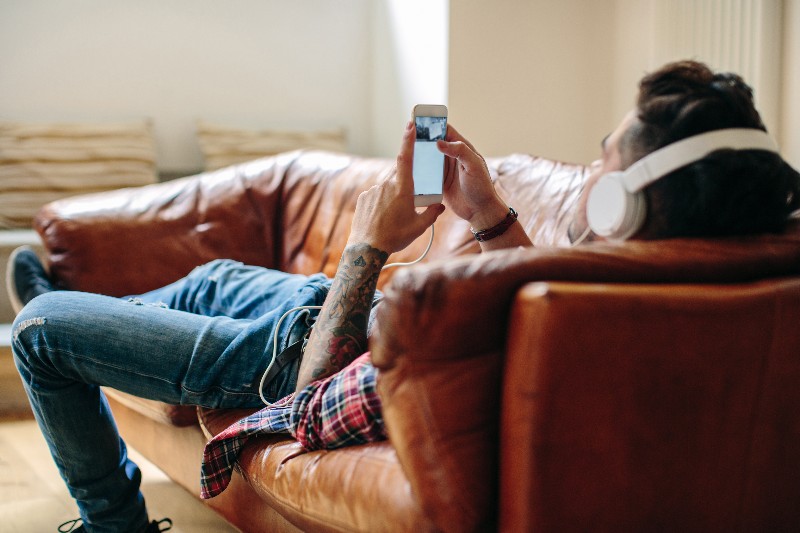 Man with white headphones lying on a couch listening to music in a flannel shirt and jeans with a tattoo sleeve on his arm