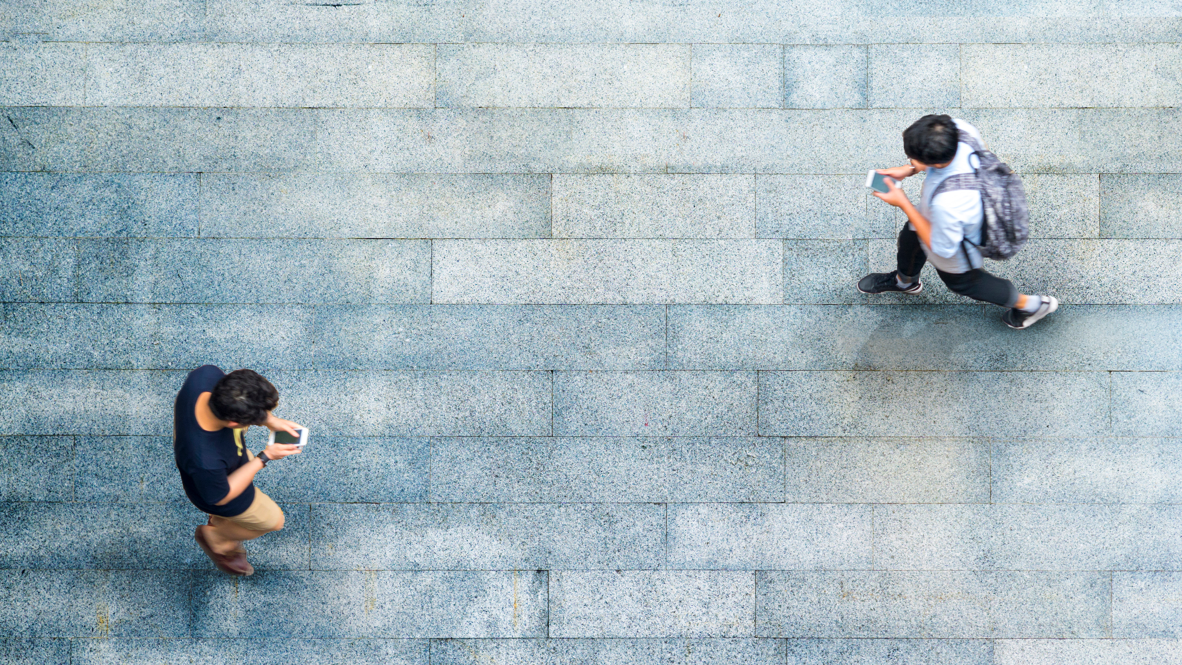 Aerial view down on two Gen Z’s males walking and looking on their mobile devices are walking towards each and about to pass