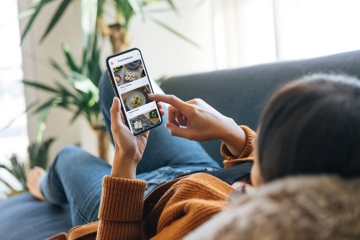 Over the shoulder view of a young woman choosing food from the menu on mobile app while lying on the couch at home.