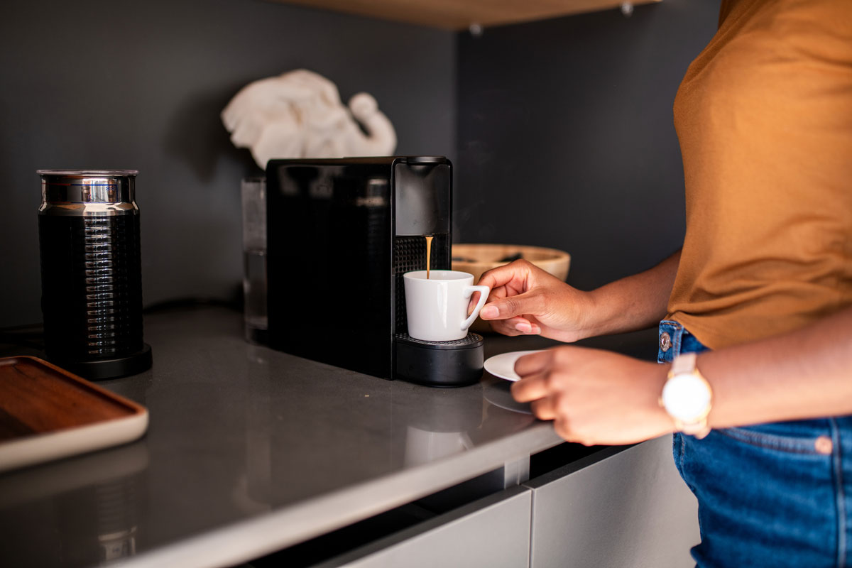 Woman in jeans and an orange shirt makes coffee in a kitchen