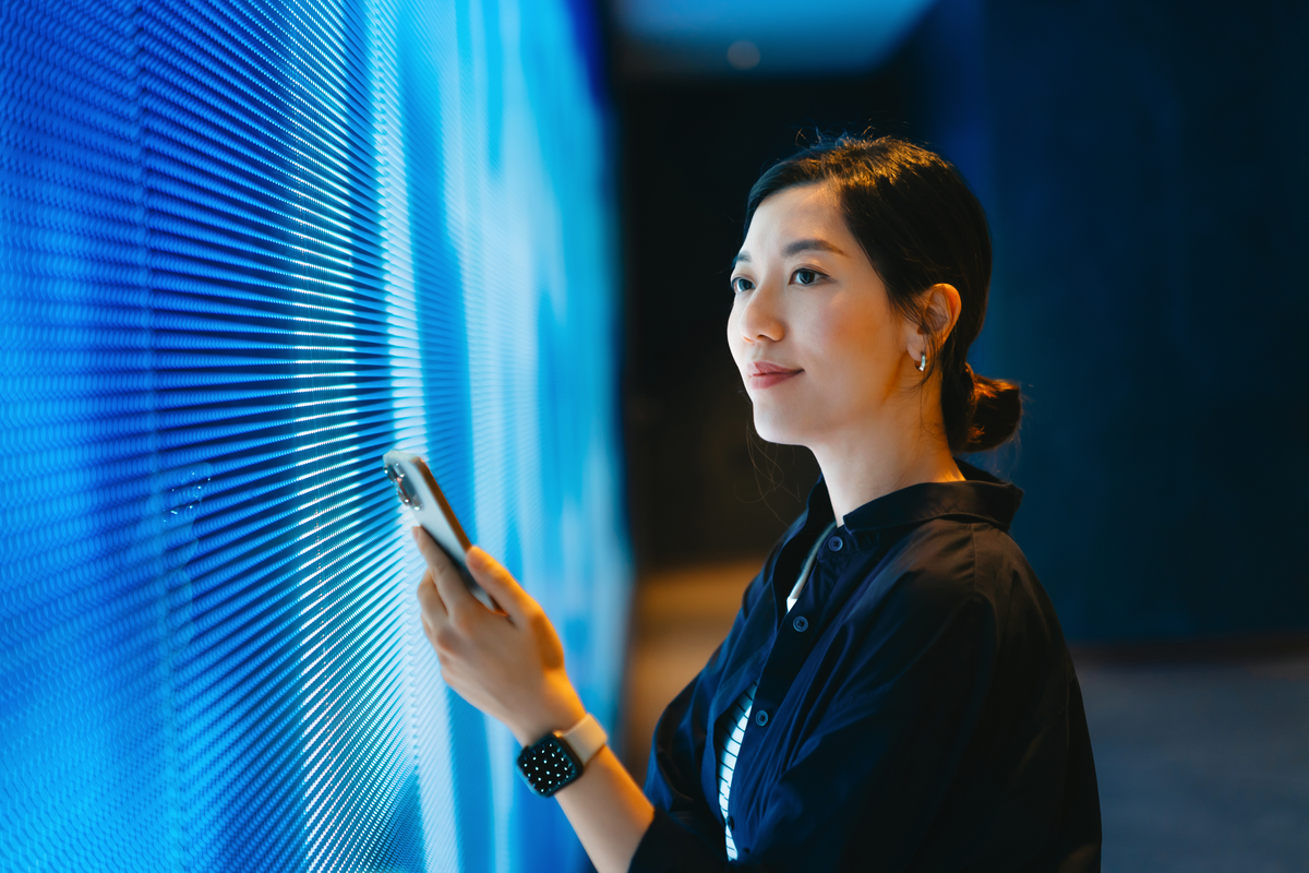 Confident young Asian woman using smartphone against blue coloured illuminated LED digital display screen in the dark. Connecting to the future. Futuristic. Artificial intelligence. Innovation, lifestyle and technology. Smart banking with technology