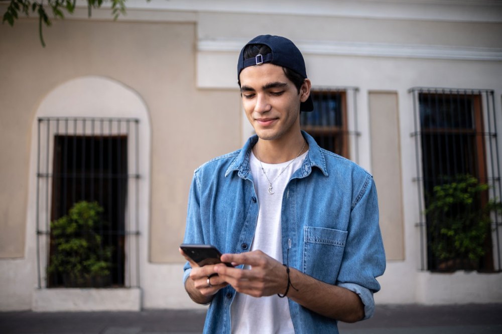 A young Latino teenager wearing a blue cap and a blue jean shirt holds his cell phone outside of a residence