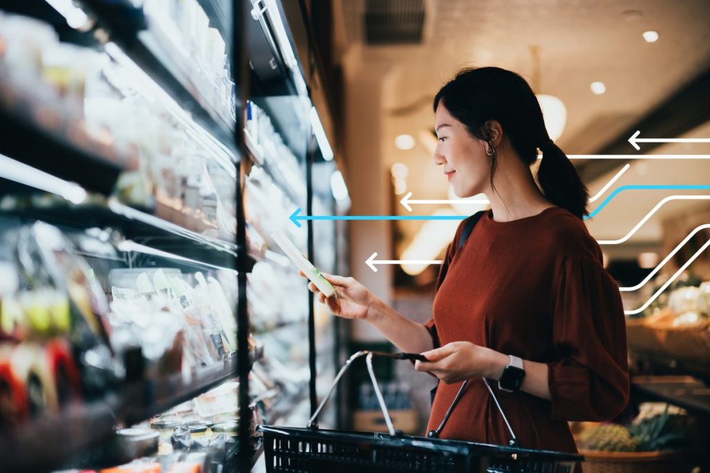 Young Asian woman carrying a shopping basket while grocery shopping in supermarket, standing along the dairy aisle, reading the nutrition label on a packet of fresh organic cheese. Making healthier food choices