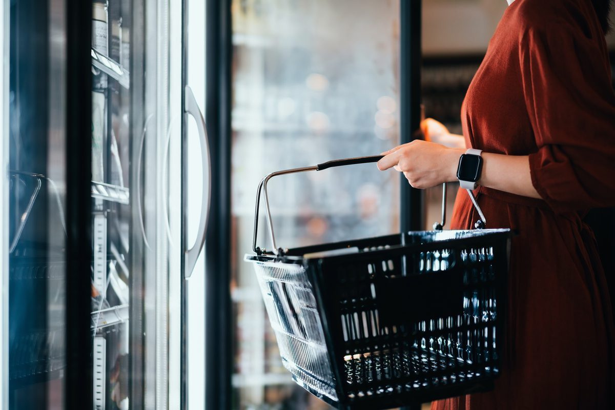 Mid-section of young Asian woman carrying a shopping basket, standing along the cold produce aisle, shopping for frozen food and daily necessities in supermarket. Healthy eating lifestyle. Making healthier food choices. Home cooking lifestyle