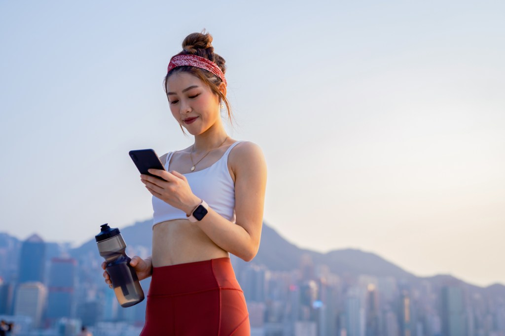 oung Asian sports woman using smartphone after exercising outdoors in city in the morning, against urban city skyline. Relaxing after running. Youth culture. Habits and hobbies. Active lifestyle. Health and fitness with technology concept