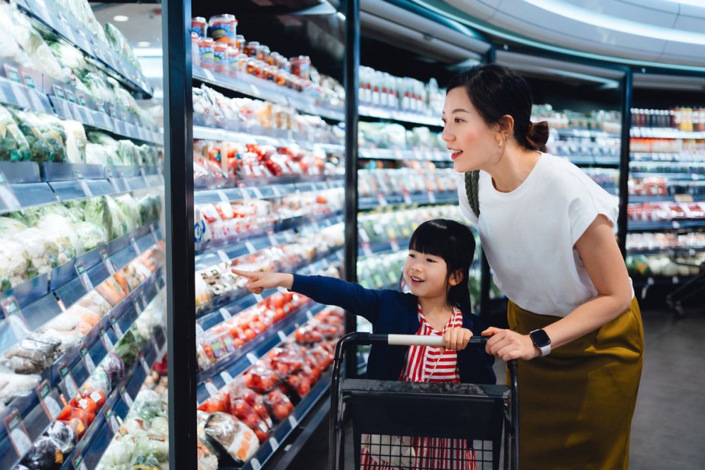 An Asian mother in a white shirt pushes a shopping cart with her daughter in it in a store
