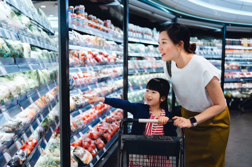 An Asian mother in a white shirt pushes a shopping cart with her daughter in it in a store