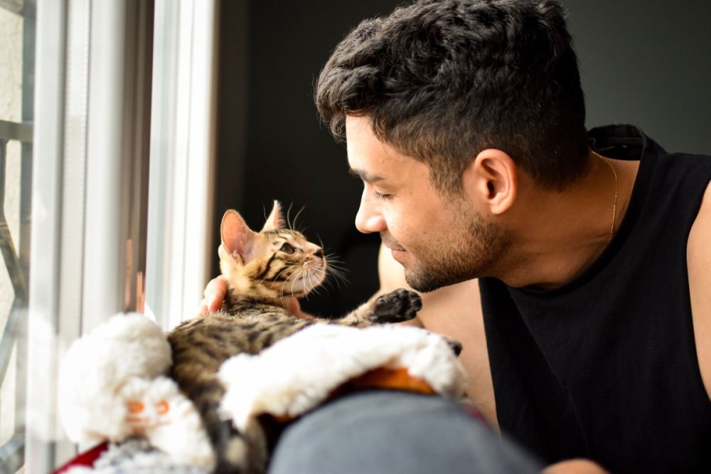 Man with dark hair and sleeveless shirt pets a cat near a window