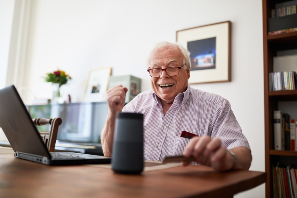Smiling retired man purchasing online using wireless devices at home. Senior man doing online shopping using a credit card, laptop and digital voice assistant.