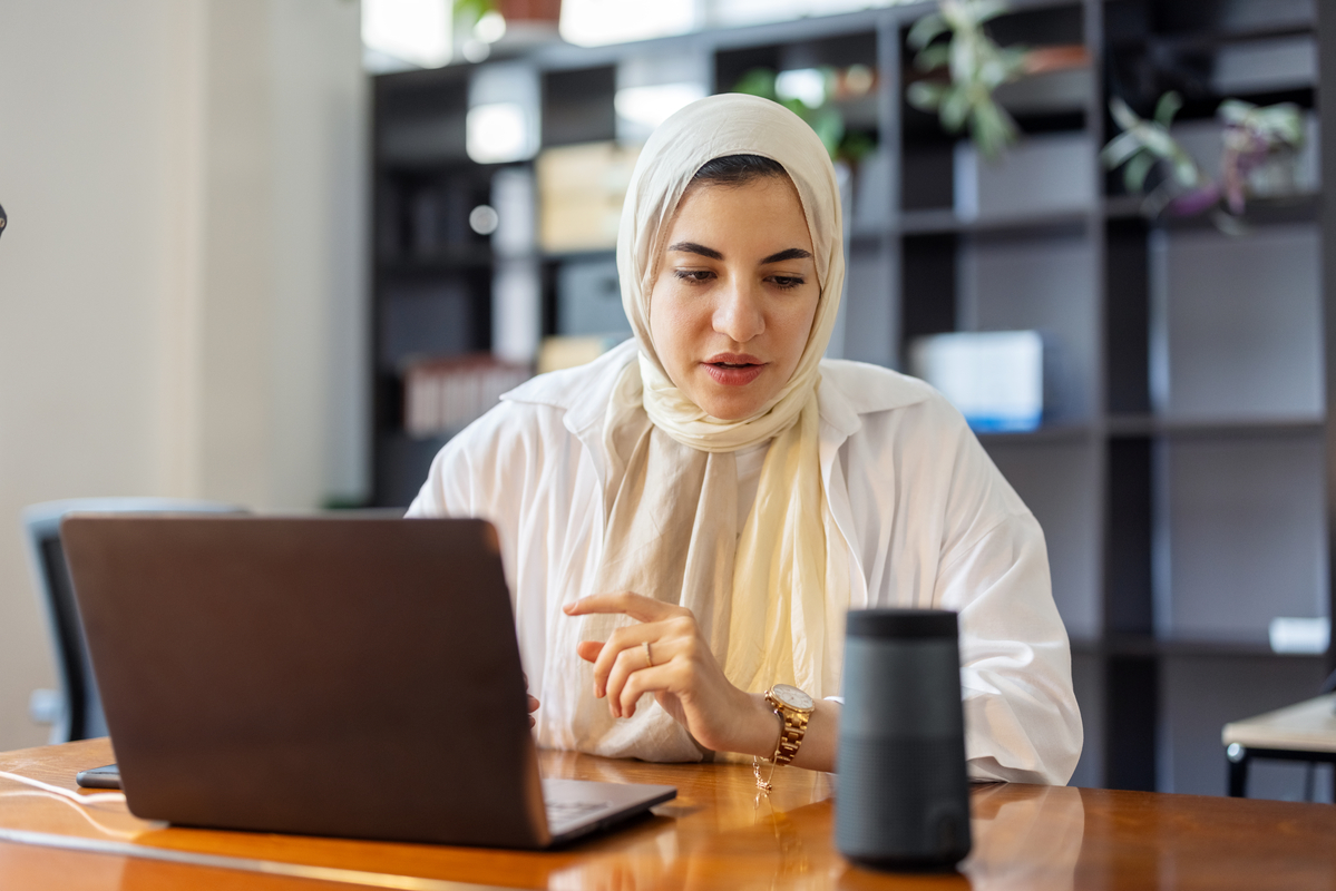 Islamic businessman using smart speaker while working at her desk in office. Woman using virtual assistant on office table while working on laptop.