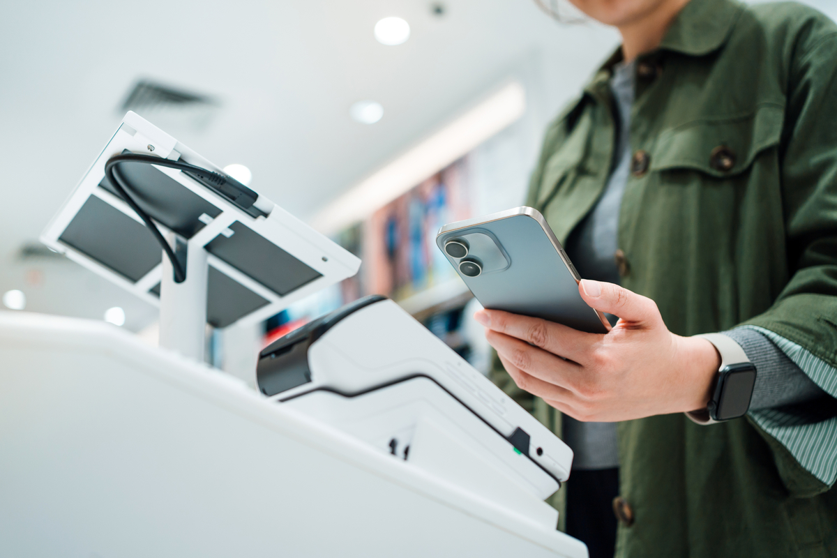Close up of a man's hand making mobile payment with her smartphone in a shop, scan and pay a bill on a card machine making a quick and easy contactless payment at self checkout counter. NFC technology, tap and go concept
